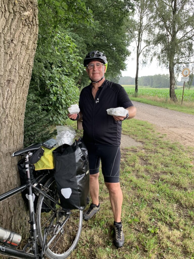 Pause in Maria Veen. David steht neben seinem Fahrrad mit zwei Brottüten in der Hand.