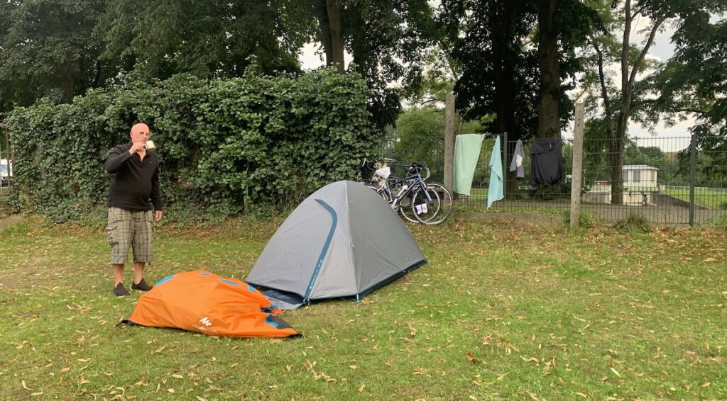 David steht mit einer Tasse Kaffee an unserem Zelt auf dem Campingplatz am Waldsee in Lingen